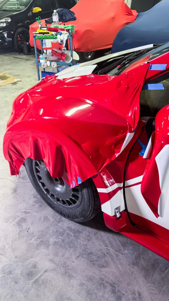 Pose d’un film adhésif rouge sur l’aile avant d’une voiture de course du Trophée Andros lors de la reproduction de son covering de décoration d’origine