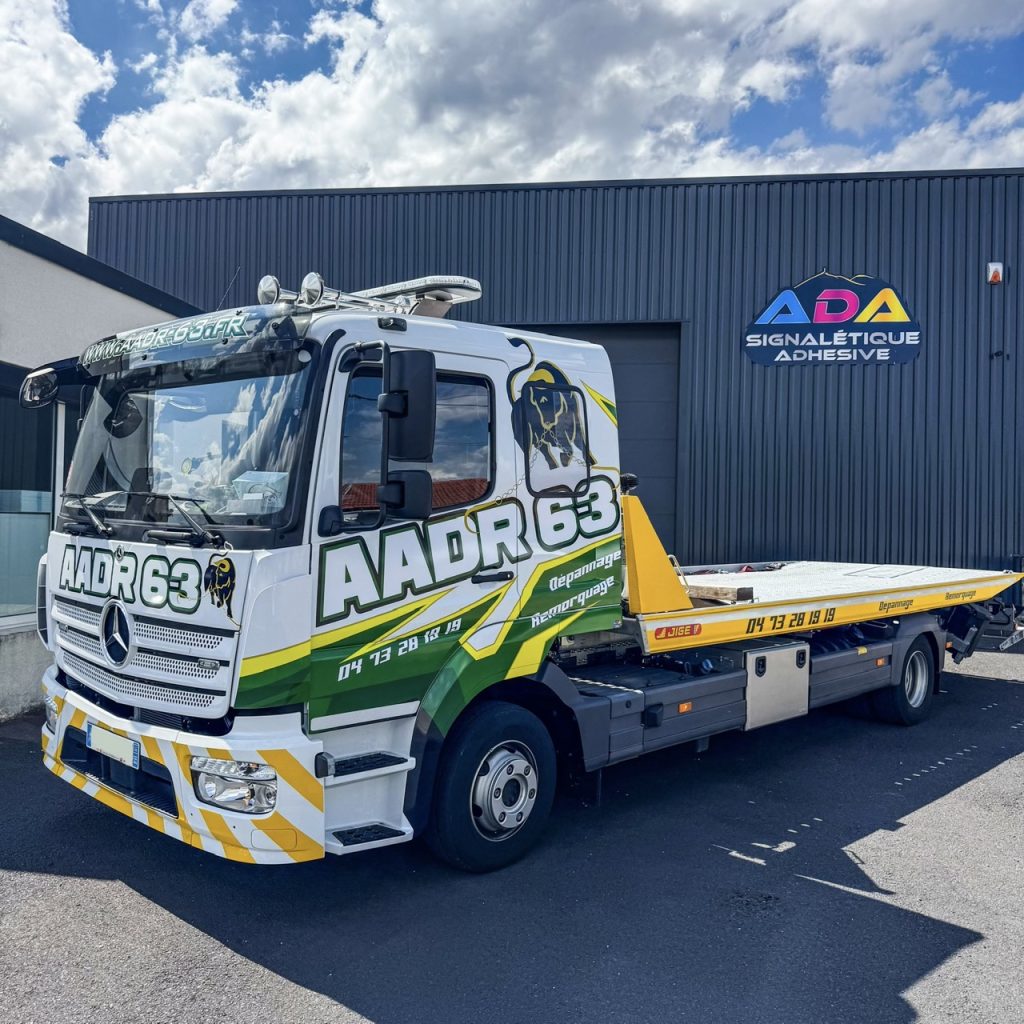 Dépanneuse Mercedes AADR 63 avec marquage publicitaire jaune, vert et blanc devant l’atelier ADA Signalétique